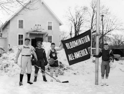 Four boys in sports uniforms standing in snow in fornt of City Hall.